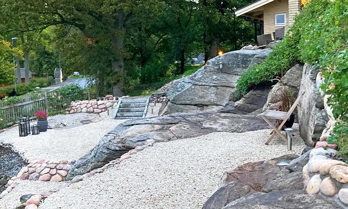 En vacker uteplats sten med grusgångar, stenmurar och en trappa som leder upp mot ett hus.