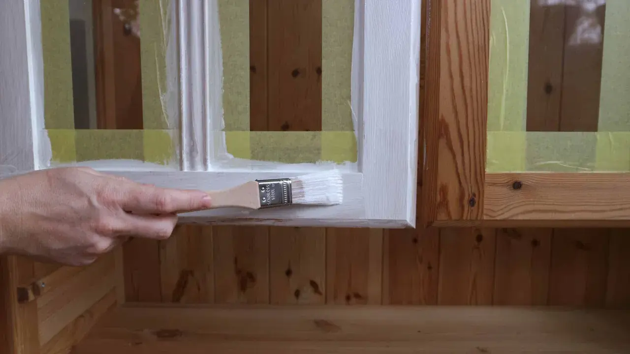 En hand målar en vit ram på en furumöbel. Maskeringstejp skyddar träet.