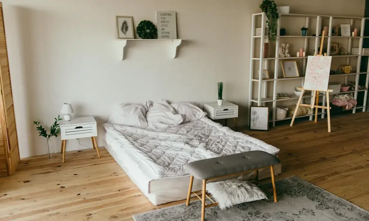 Ett sovrum med en stor, vit madrass som ser ut att vara förvaring under. En hylla med dekorationer, ett staffli med en tavla och en bokhylla fyller rummet.