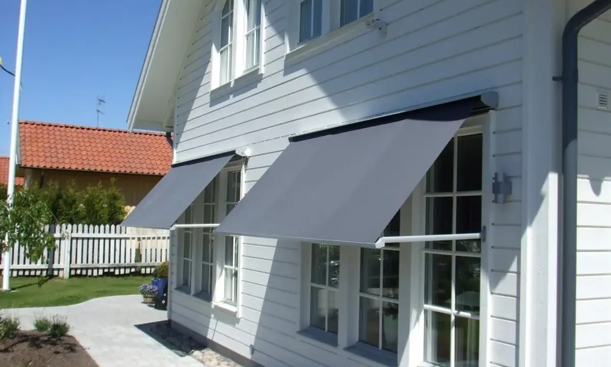 Grå markiser monterade på ett vitt trähus. Solen skiner och skapar skuggor på fasaden.
