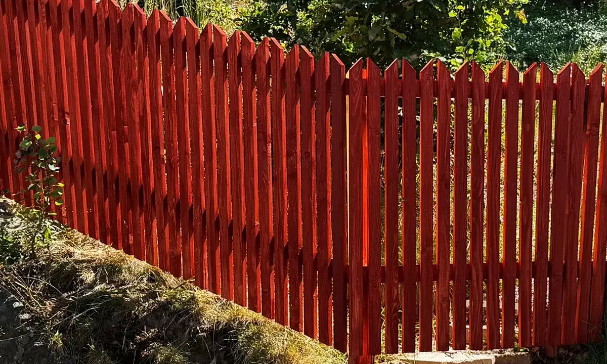 Rött, spetsigt staket av tryckimpregnerat trä. Solen lyser genom plankorna och skapar skuggor.