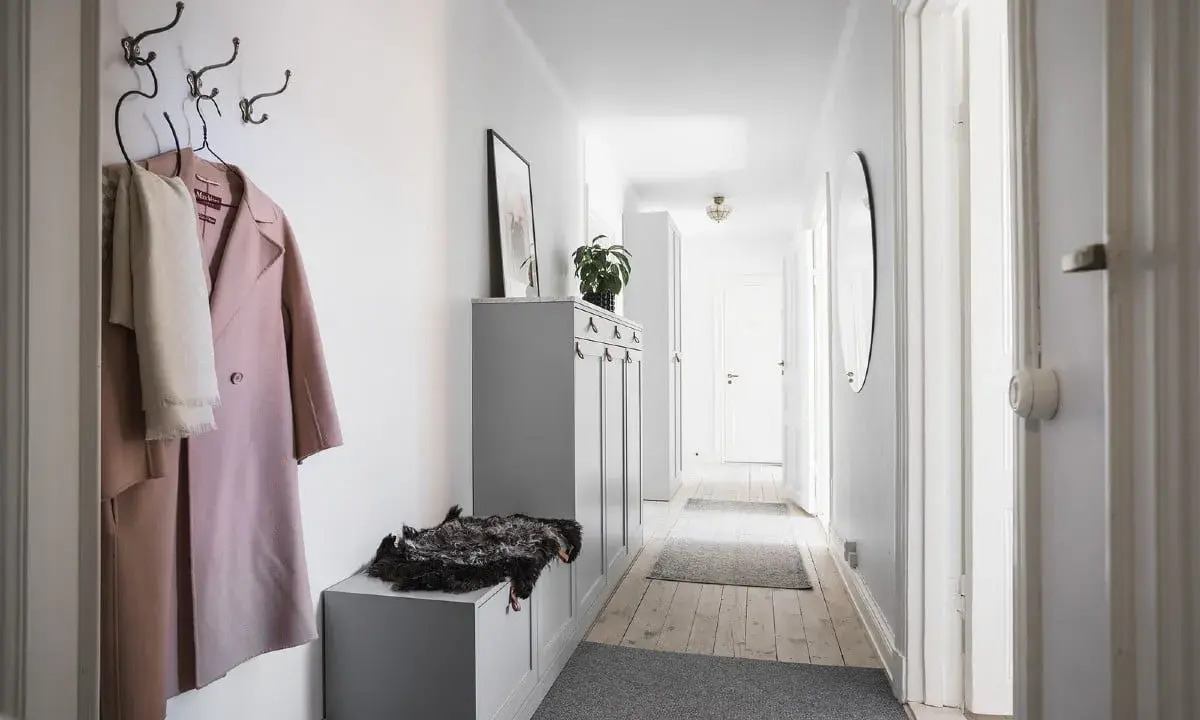 En smal hall med en rosa kappa och en vit halsduk på krokar. En grå förvaringsmöbel med en fårskinnspläd.