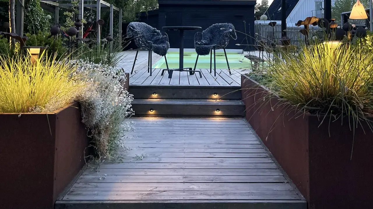 Välkomnande altan med decklights som lyser upp trappstegen. Två mysiga fåtöljer väntar vid ett runt bord.