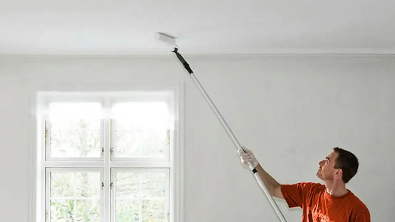 Man målar taket med en lång roller. Fönstret bakom släpper in ljus.