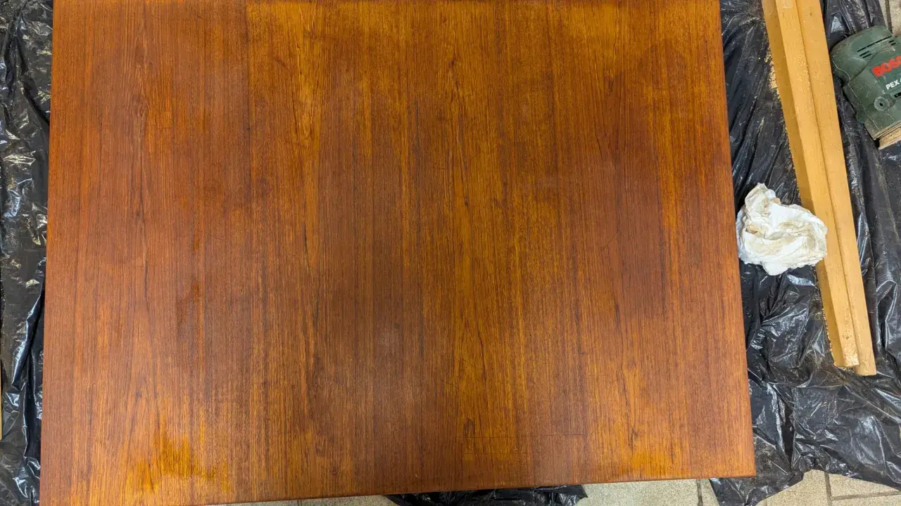 En träbordsskiva ligger på svart plast. Bredvid ligger en slipmaskin och en trasa, redo att måla bordsskivan.