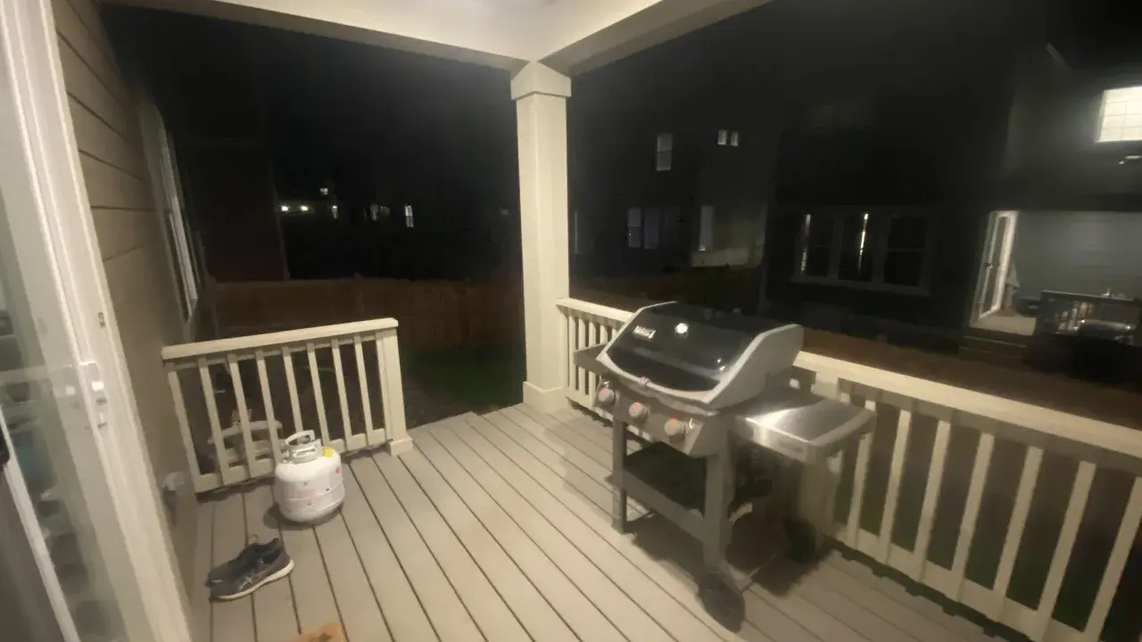 En gasolgrill under taket på altanen, redo för grillkvällar. Bredvid står en gasoltub och ett par skor.