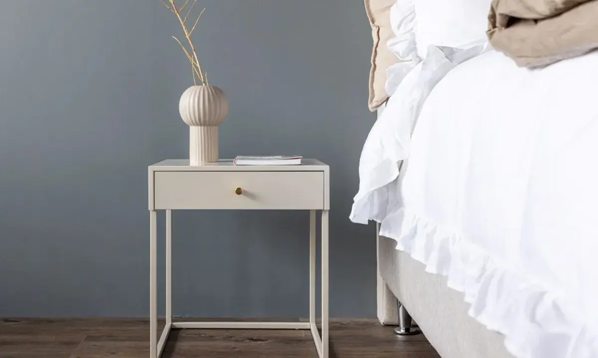 Ett stilrent sängbord i beige med en låda och en vas med kvistar. Bredvid står en inbjudande säng med vitt, volangprytt överkast.