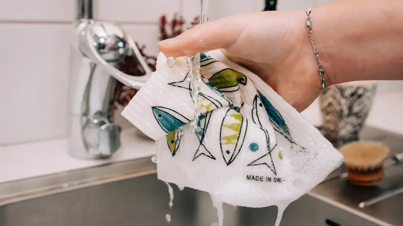 En hand håller en disktrasa i linne med fiskmönster under rinnande vatten i ett kök.
