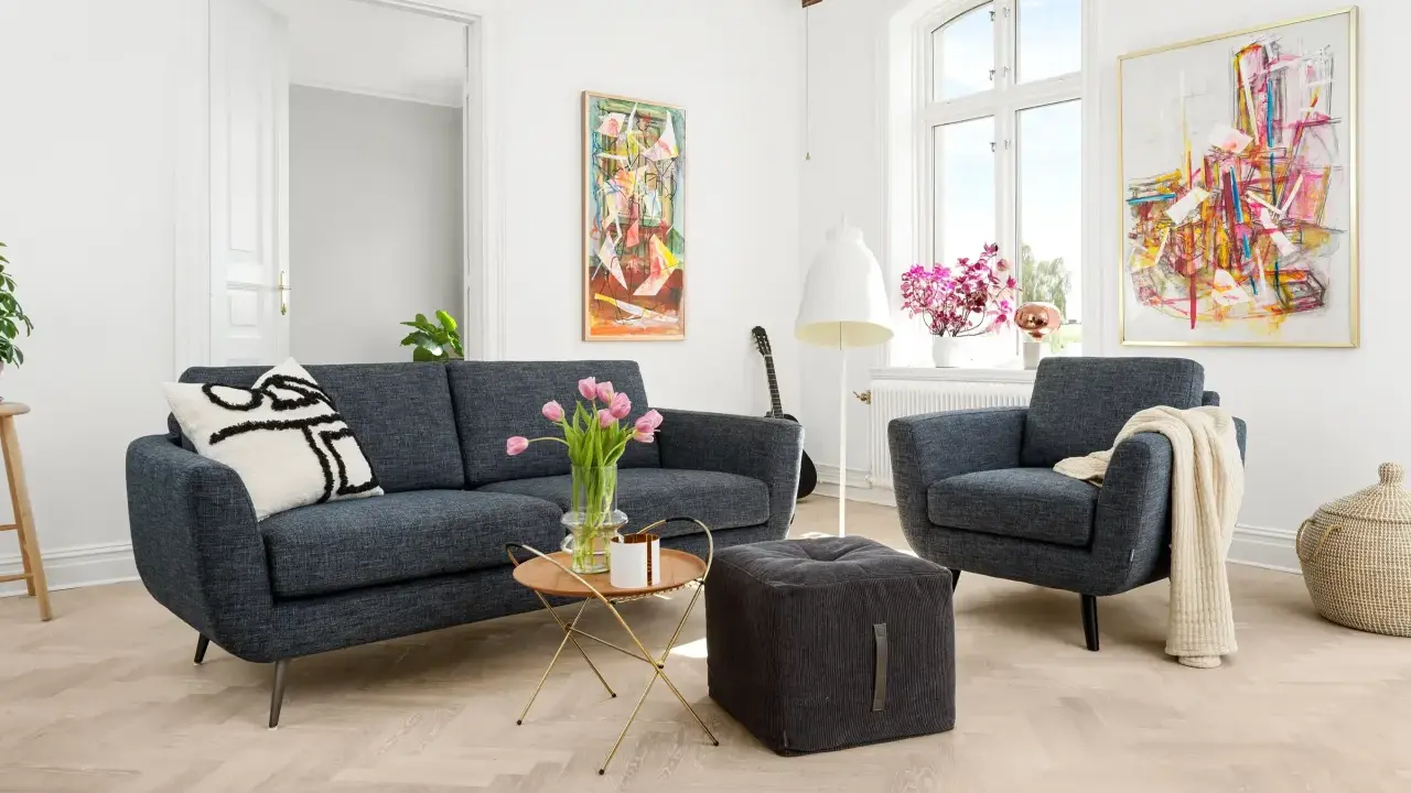 En modern soffgrupp 3+2 i mörkblått tyg, med en fåtölj, soffbord, pall och konst på väggarna.