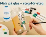 En hand målar ett blommönster på ett glas med en pensel. Färger och alkohol på bordet.