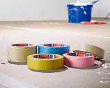 Fem rullar maskeringstejp i olika färger ligger på ett golv som täckts med papper. En blå hink och en röd roller syns i bakgrunden.