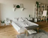 Ett sovrum med en stor, vit madrass som ser ut att vara förvaring under. En hylla med dekorationer, ett staffli med en tavla och en bokhylla fyller rummet.