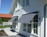 Grå markiser monterade på ett vitt trähus. Solen skiner och skapar skuggor på fasaden.
