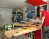Man i röd tröja i en inredd verkstad. Verktyg på väggen, bänkar med maskiner och material.
