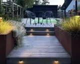 Välkomnande altan med decklights som lyser upp trappstegen. Två mysiga fåtöljer väntar vid ett runt bord.