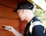 Kvinna med keps mäter luftfuktighet i en röd timmervägg med en fuktmätare.