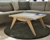 Ett smart soffbord i ljust trä med en vit inläggning, placerat på en rund, mörk matta framför en beige hörnsoffa.