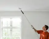 Man målar taket med en lång roller. Fönstret bakom släpper in ljus.
