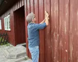 Man slipar träfasaden på ett rött hus. Förberedelser för att måla om.