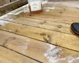 En flaska linoljesåpa står på en altan som håller på att rengöras. En svart stövel med orange kant syns i bildens nedre högra hörn.
