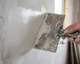 En hand med spackelspade breddar vit fogmassa på en vägg. Förberedelse för att måla direkt på gips.
