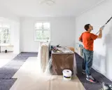 Man målar väggar, har tagit bort eluttag vid målning. Skyddsplast och färgburkar syns.