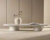 Ett modernt, ovalt soffbord med en vit puff-liknande bas. En vas med en gren och en öppen bok står på bordet.