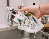 En hand håller en disktrasa i linne med fiskmönster under rinnande vatten i ett kök.