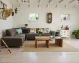 Mysigt vardagsrum med rustik möbler, mörkbrun soffa, träbord och golvlampa. Vita tegelväggar och ljusa trägolv.