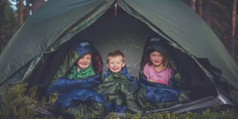 Tre glada barn myser i sina sovsäckar i ett tält. En perfekt tältsäng barn för äventyr i skogen.