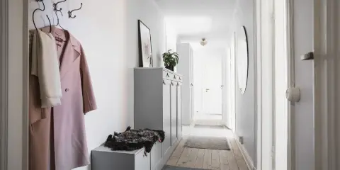 En smal hall med en rosa kappa och en vit halsduk på krokar. En grå förvaringsmöbel med en fårskinnspläd.