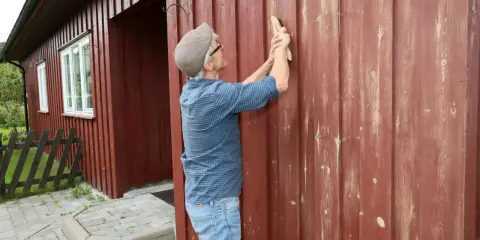 Man slipar träfasaden på ett rött hus. Förberedelser för att måla om.