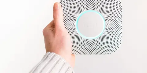 En hand håller upp en vit Google Nest Protect-rökdetektor med blåaktig glöd.