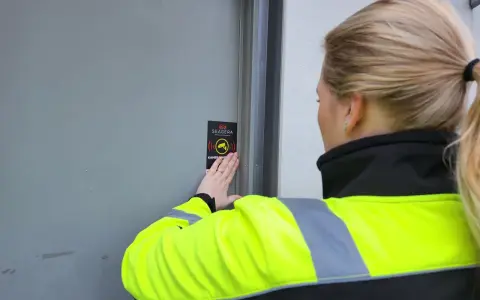 En person med reflexväst fäster en skylt med texten "Kameraövervakning" på en dörr, vilket indikerar ett larm på en byggarbetsplats.