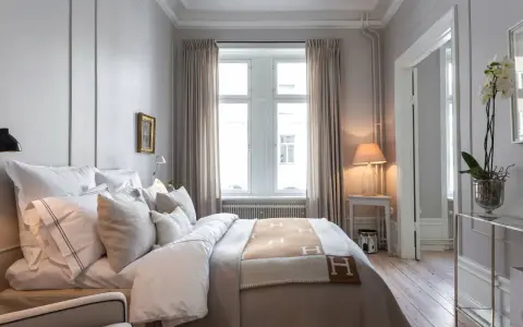 Ett elegant sovrum med en stor säng bäddad med vita lakan och en brun pläd med H-mönster. Vackra möbler till sovrummet som ett litet sidobord med lampa och en orkidé.