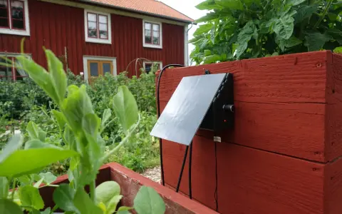 En solcellsdriven bevattning är monterad på en röd träkista i en trädgård, med ett rött hus i bakgrunden.