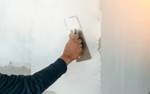 Handen sprider spackel på en vägg med en bredspackel. Arbetet med att måla på spackel fortsätter för att skapa en jämn yta.