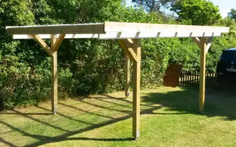 En nybyggd carport på plintar står på en gräsmatta. Träkonstruktionen har ett tak av plast eller metall.