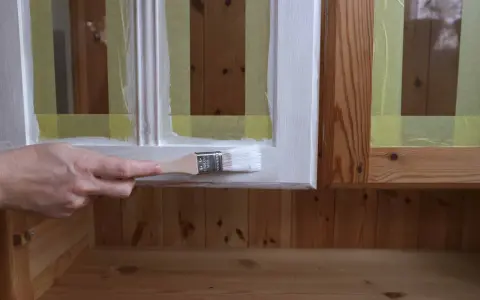 En hand målar en vit ram på en furumöbel. Maskeringstejp skyddar träet.