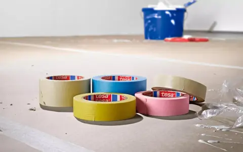 Fem rullar maskeringstejp i olika färger ligger på ett golv som täckts med papper. En blå hink och en röd roller syns i bakgrunden.