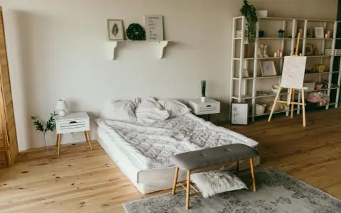 Ett sovrum med en stor, vit madrass som ser ut att vara förvaring under. En hylla med dekorationer, ett staffli med en tavla och en bokhylla fyller rummet.