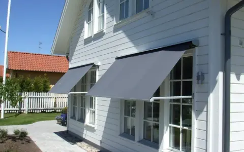 Grå markiser monterade på ett vitt trähus. Solen skiner och skapar skuggor på fasaden.
