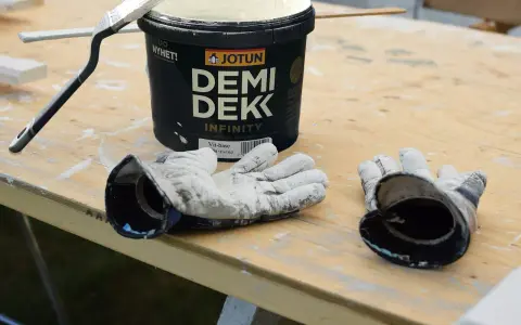 Förberedelser för målning utomhus: en burk Jotun Demi Dekk, pensel och slitna handskar ligger på ett träbord.
