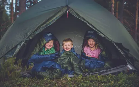 Tre glada barn myser i sina sovsäckar i ett tält. En perfekt tältsäng barn för äventyr i skogen.