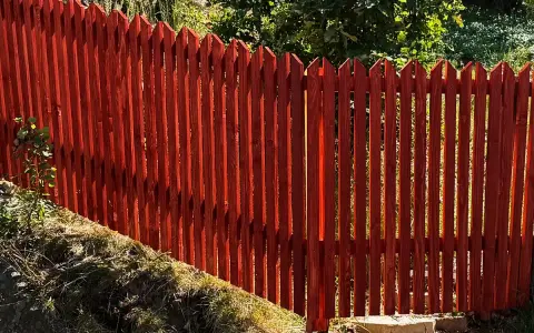 Rött, spetsigt staket av tryckimpregnerat trä. Solen lyser genom plankorna och skapar skuggor.