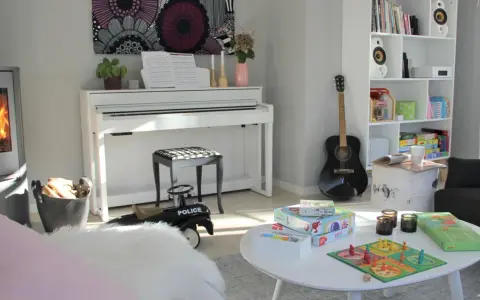 Ett mysigt matrum med en ståtlig kamin, ett vitt piano och en bokhylla fylld med böcker och leksaker.
