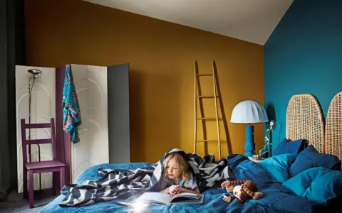 Ett barn läser under ett mönstrat täcke i ett mysigt blått sovrum. En golvlampa och en stege i trä bidrar till rummets unika atmosfär.