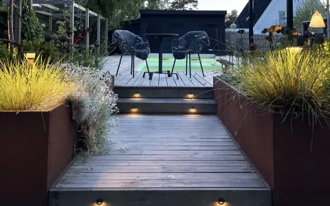 Välkomnande altan med decklights som lyser upp trappstegen. Två mysiga fåtöljer väntar vid ett runt bord.