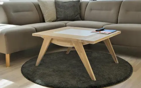 Ett smart soffbord i ljust trä med en vit inläggning, placerat på en rund, mörk matta framför en beige hörnsoffa.
