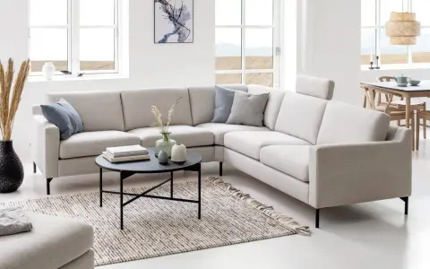 Ljusbeige hörnsoffor i ett modernt vardagsrum med ett runt soffbord, en matta och stora fönster.